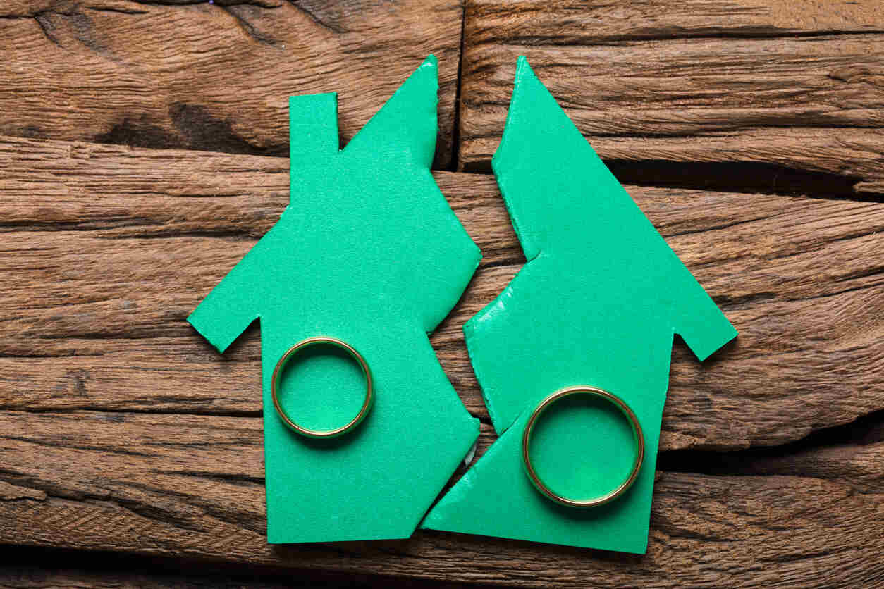 
A paper cutout of a house torn in half, with a wedding ring on each piece, sits on a wooden surface. This image represents the challenge of valuing and dividing real estate and other assets in a high-stakes California divorce.