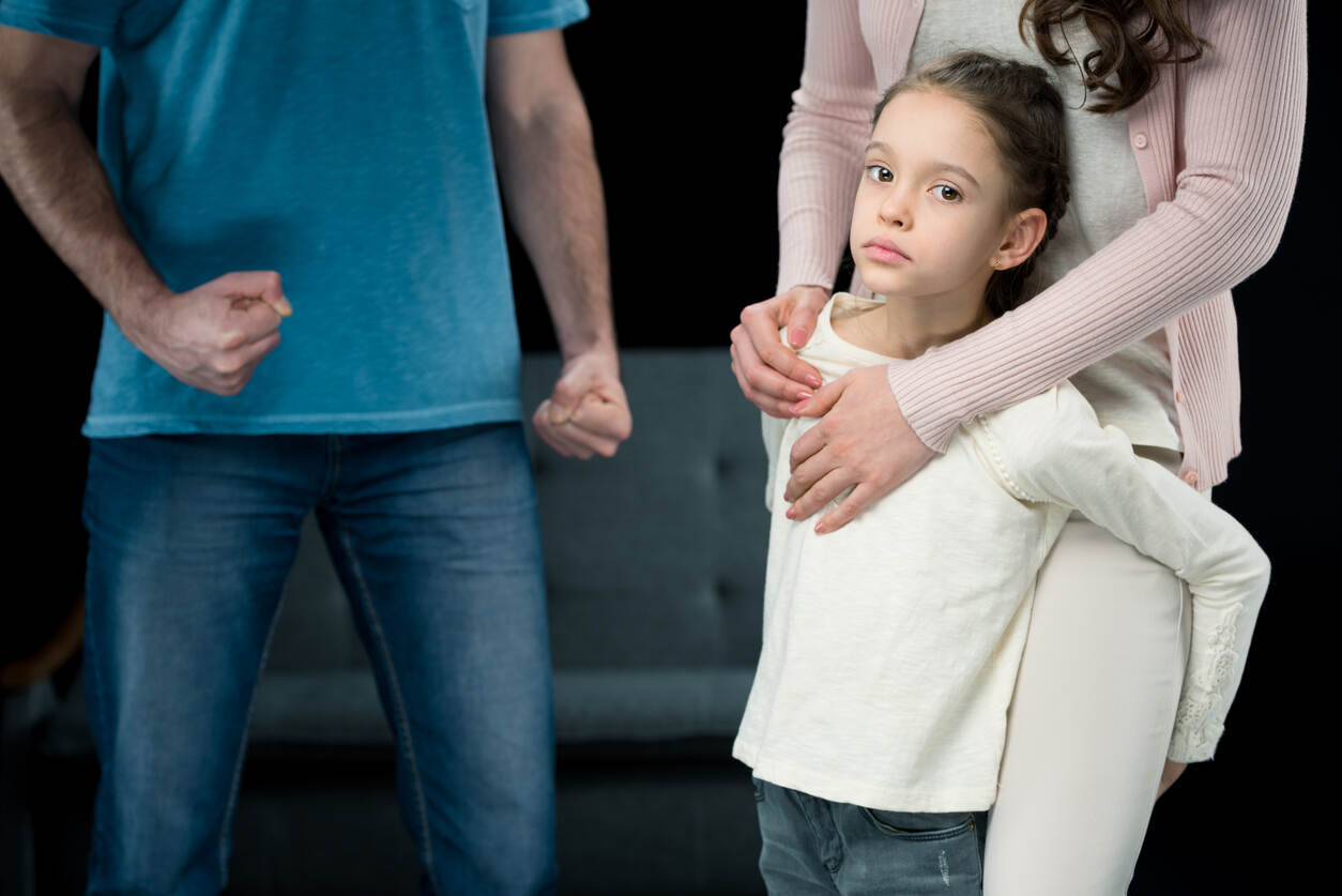 A tense family dynamic is depicted with a mother holding her daughter close, as the father stands in the background with his fists clenched. The daughter looks directly at the camera with a sad and worried expression. The image represents the emotional turmoil and conflict involved in contentious child custody cases and relocation requests in California